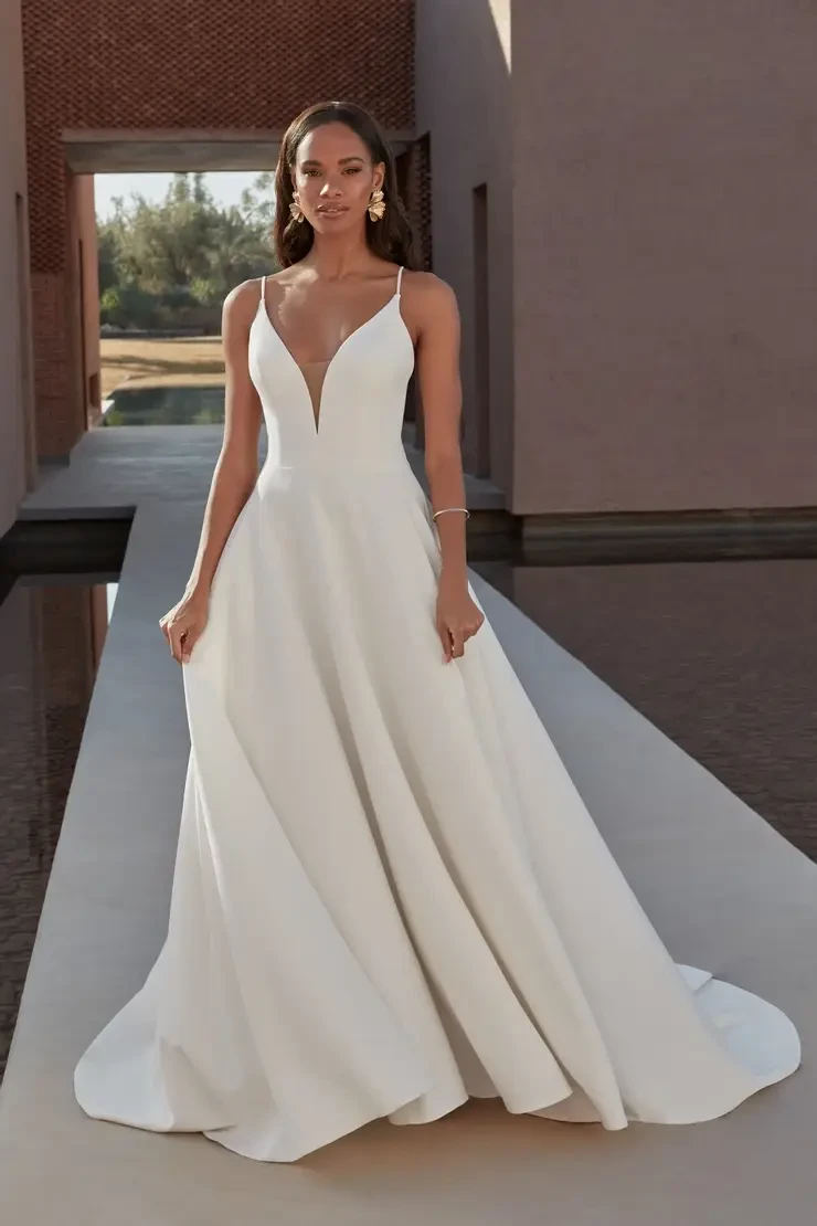 A woman in an elegant white wedding dress with thin straps and a deep V-neckline stands confidently on a reflective water pathway, framed by modern architecture.