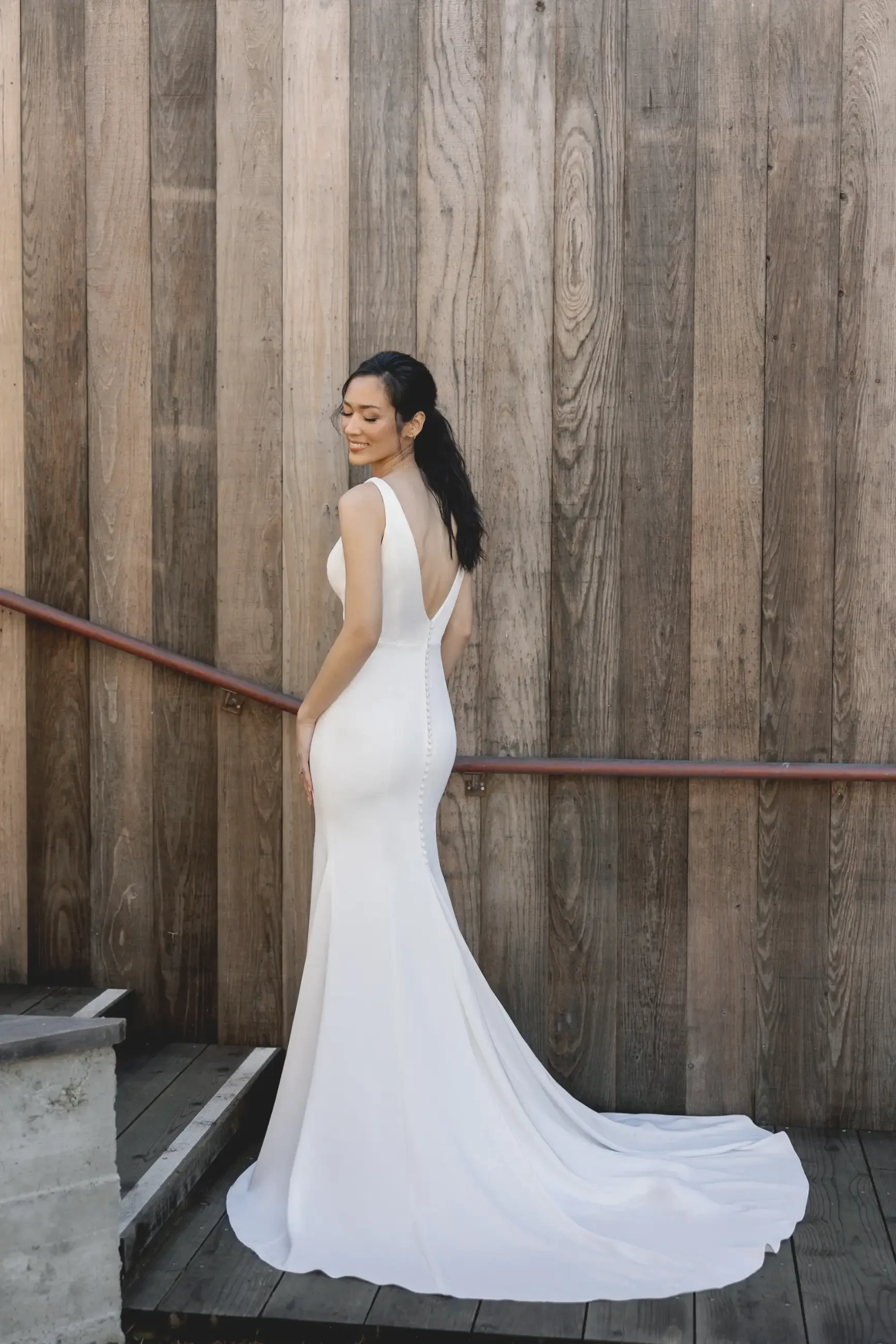 A woman in an elegant white gown stands gracefully on wooden steps. The long train of the dress flows behind her. She looks over her shoulder, exuding confidence.