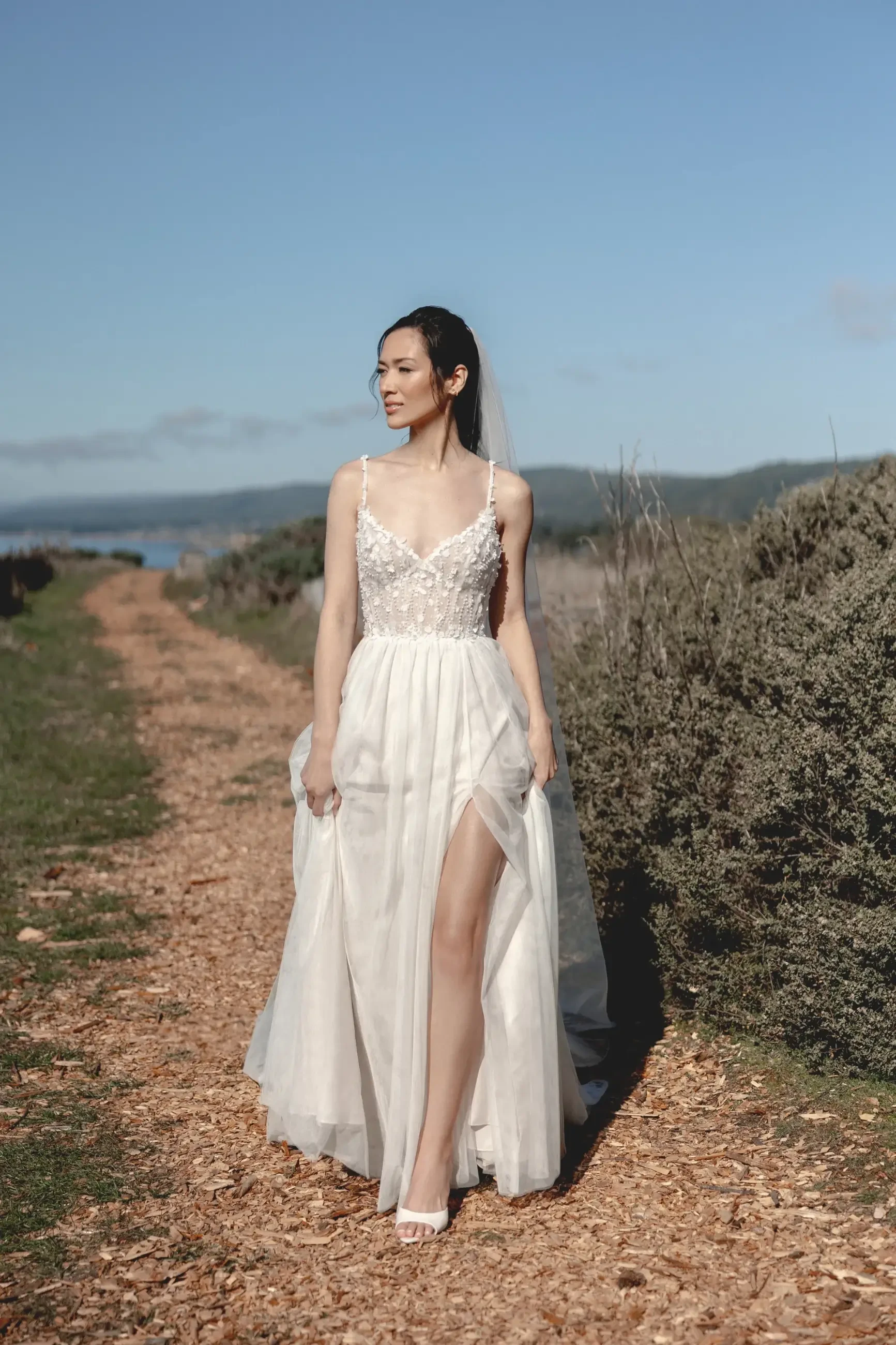A bride in a flowing white wedding dress with lace details and a thigh-high slit walks along a rustic path. Surrounding her are green shrubs, under a clear blue sky.