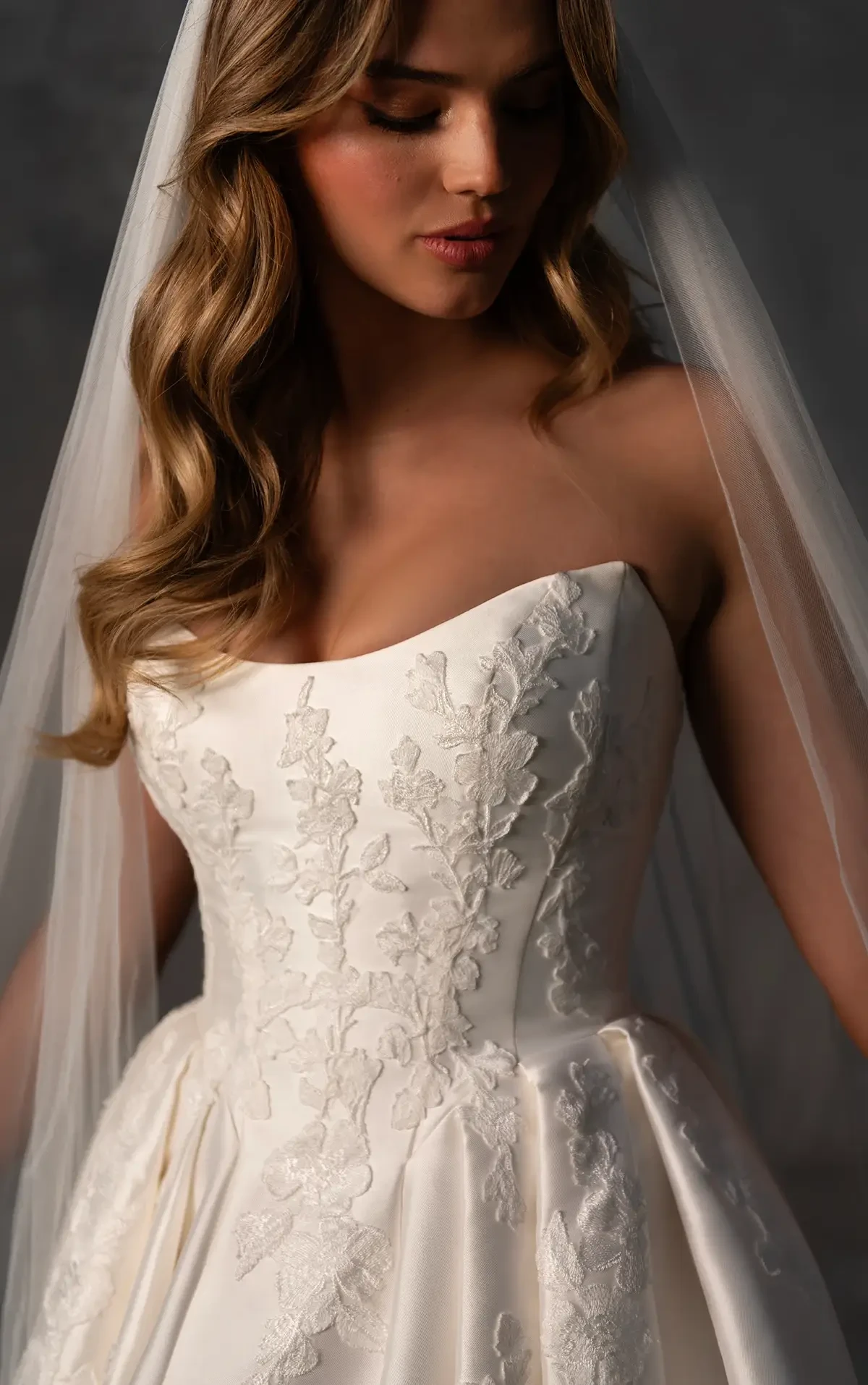 Model is wearing an Essense of Australia #D4265 gown, a strapless Mikado ballgown with a scooped cat-eye neckline, sculpted pleats, voluminous skirt, and fabric-covered button detailing along the dramatic train.