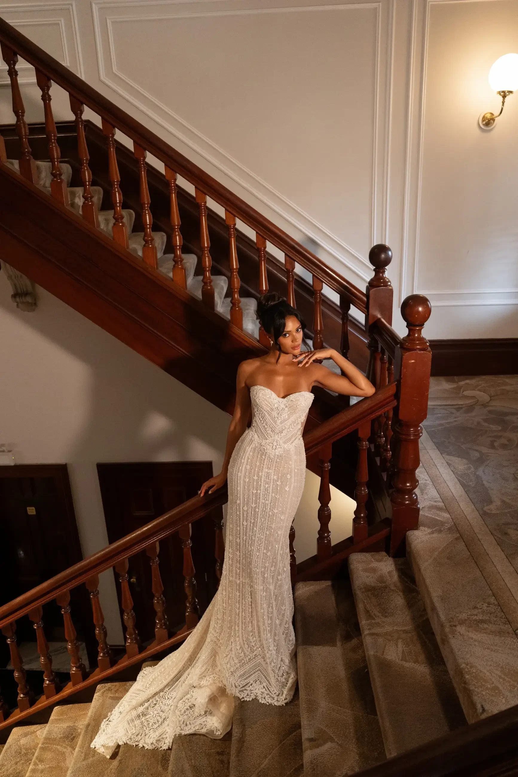 Woman in an elegant strapless white gown poses on a wooden staircase. The dim lighting and classic decor create a sophisticated, timeless ambiance.