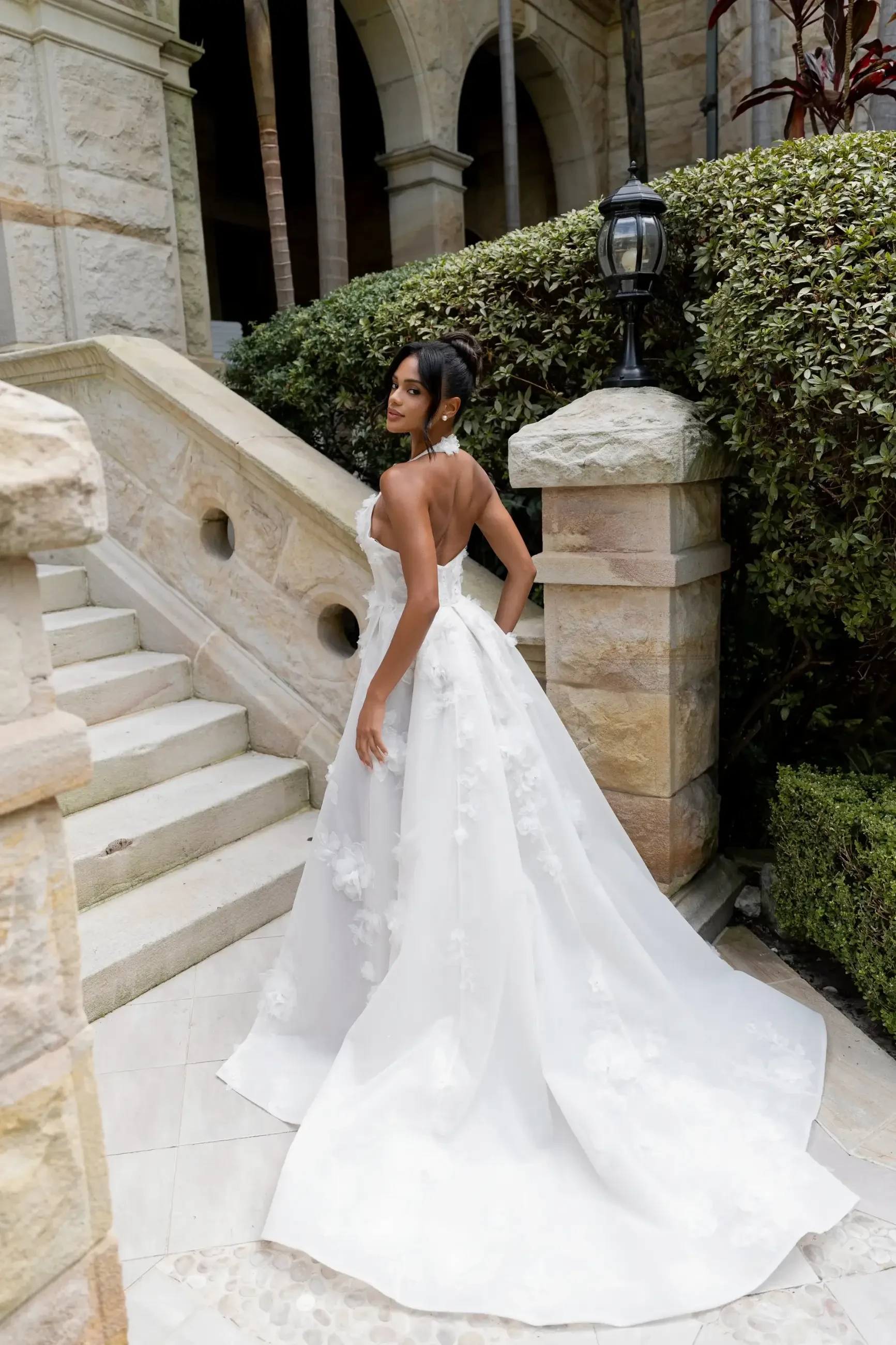 Bride in elegant white gown stands on stone path, glancing back. Surrounded by lush greenery and an ornate stone staircase, conveying romance.