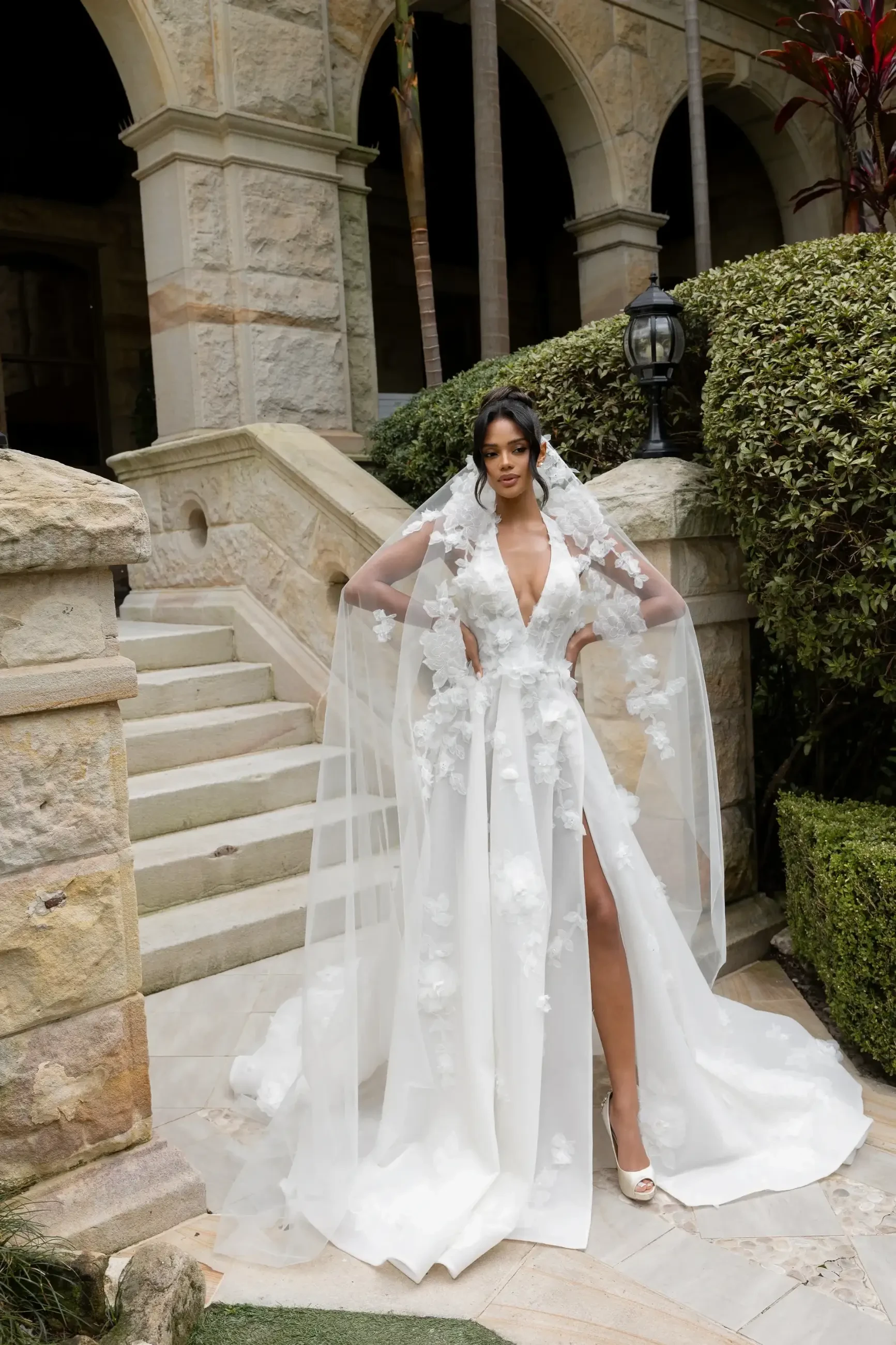 Woman in an elegant white floral gown and sheer cape stands confidently near stone steps, surrounded by lush greenery and ornate architecture. Luxurious, serene setting.