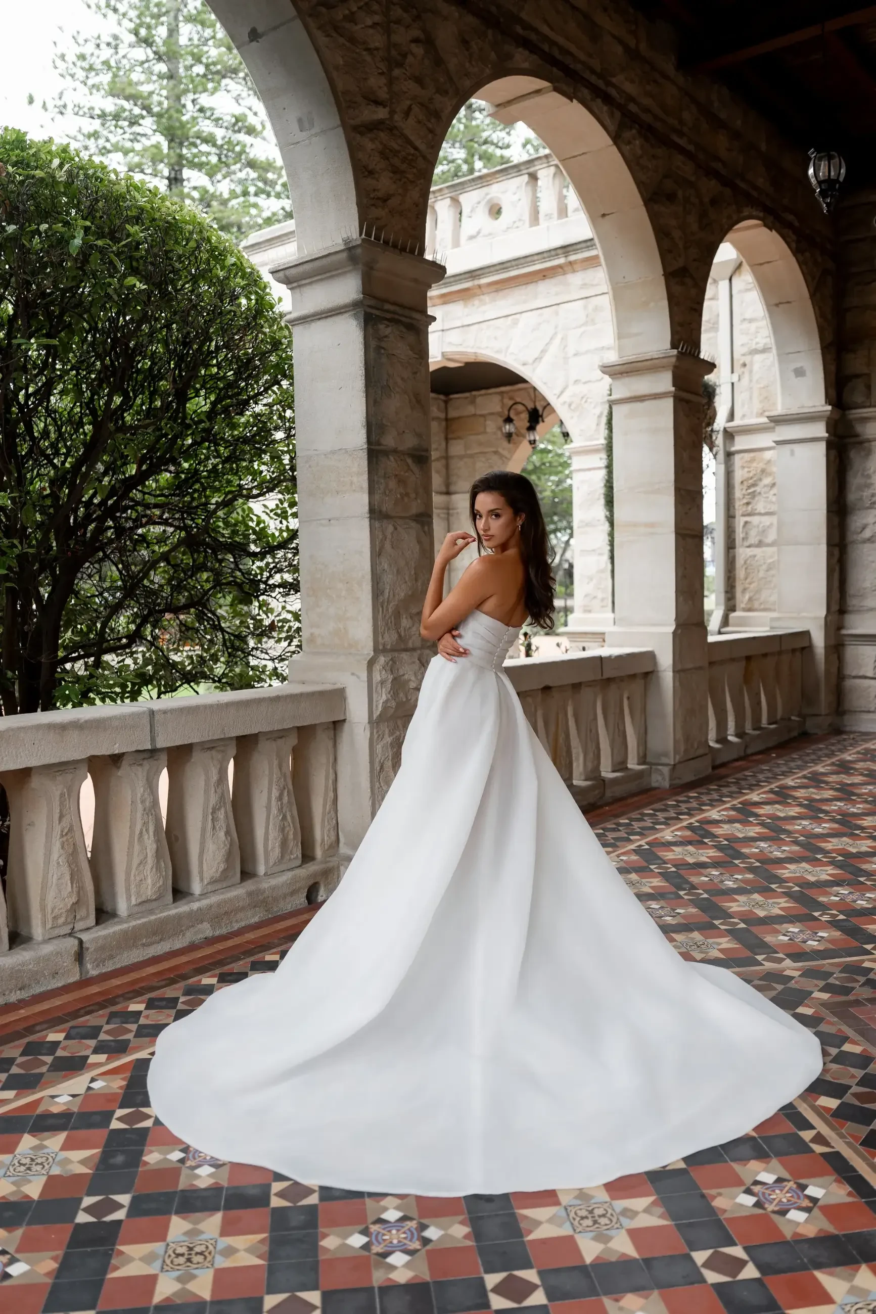 Bride in a strapless white gown stands on a patterned tile floor in an arched stone corridor, surrounded by greenery, exuding elegance and poise.