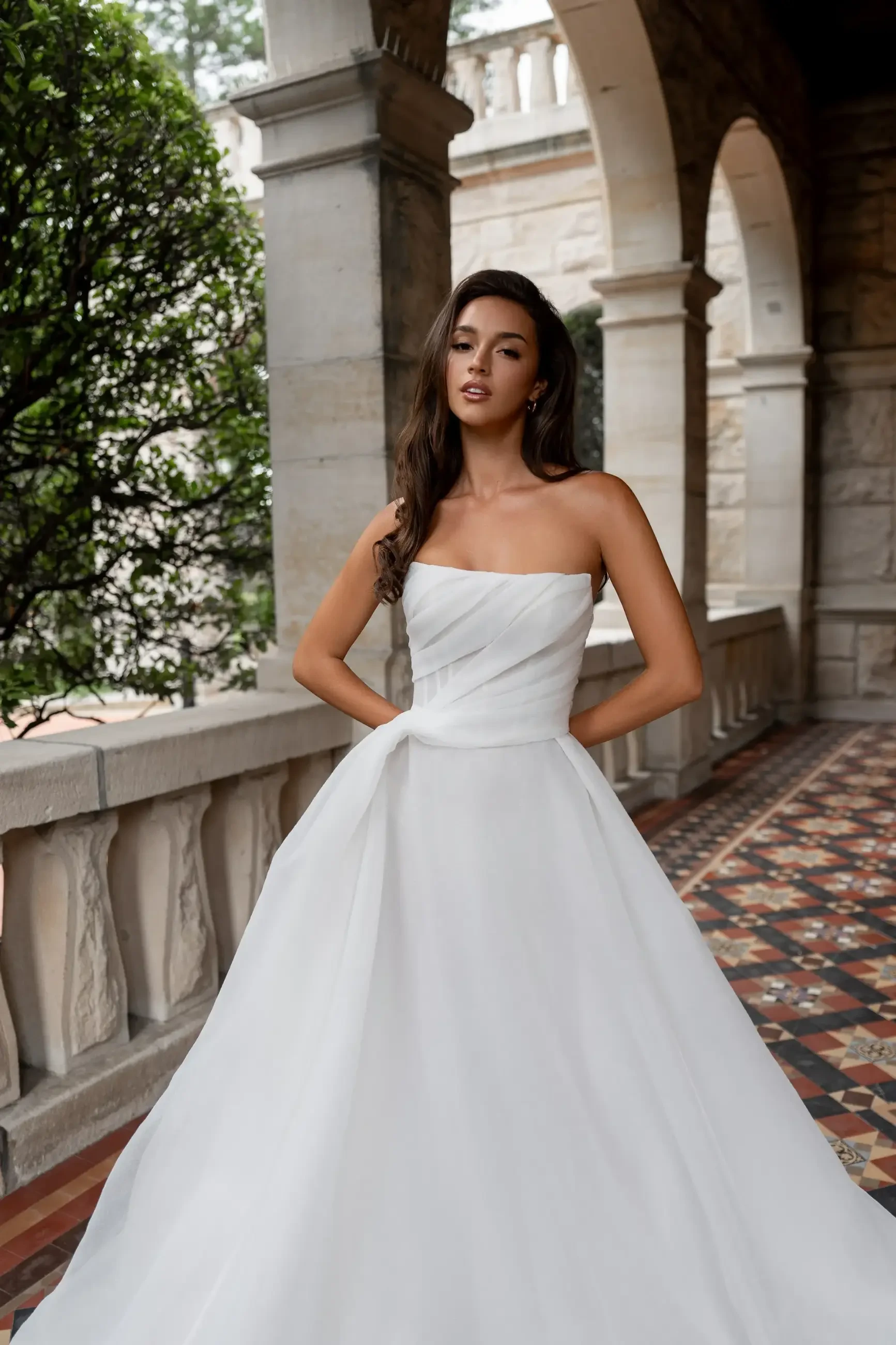 A woman in a strapless white wedding gown poses confidently in a stone corridor. The floor has a patterned design, and trees are visible outside.