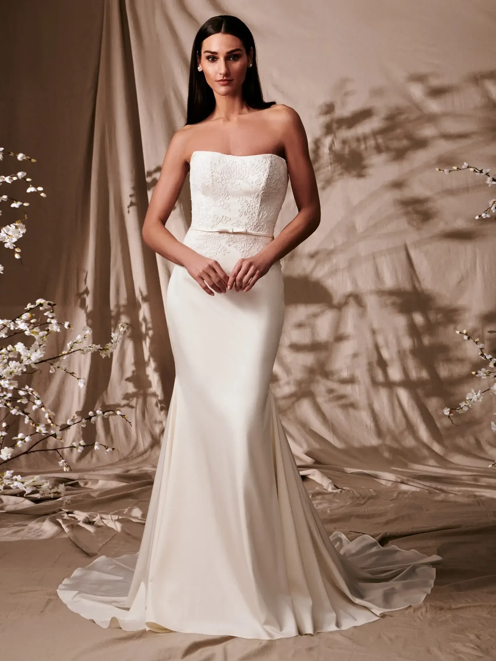 Model is wearing a fit-to-flare lace gown with removable puff sleeves, a satin bow belt, and a detachable oversized bow with streamers at the back.