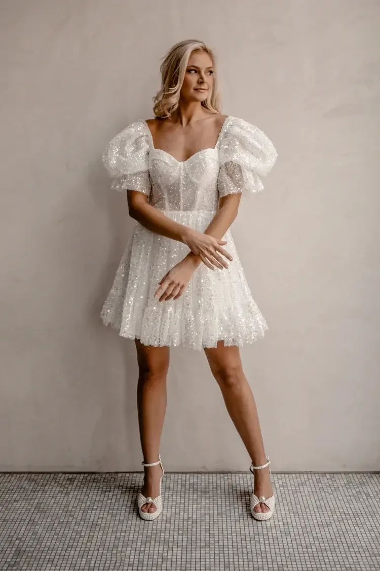 A woman is sitting on a brown couch, wearing a sparkly white off-the-shoulder dress and white high-heeled shoes.