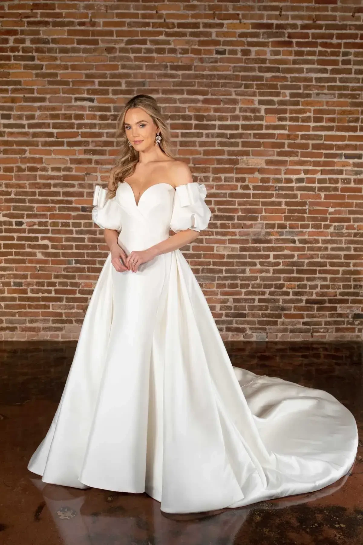 A woman in an elegant white off-shoulder wedding gown with puff sleeves stands on a polished floor, backed by a rustic brick wall. She exudes a serene, confident demeanor.