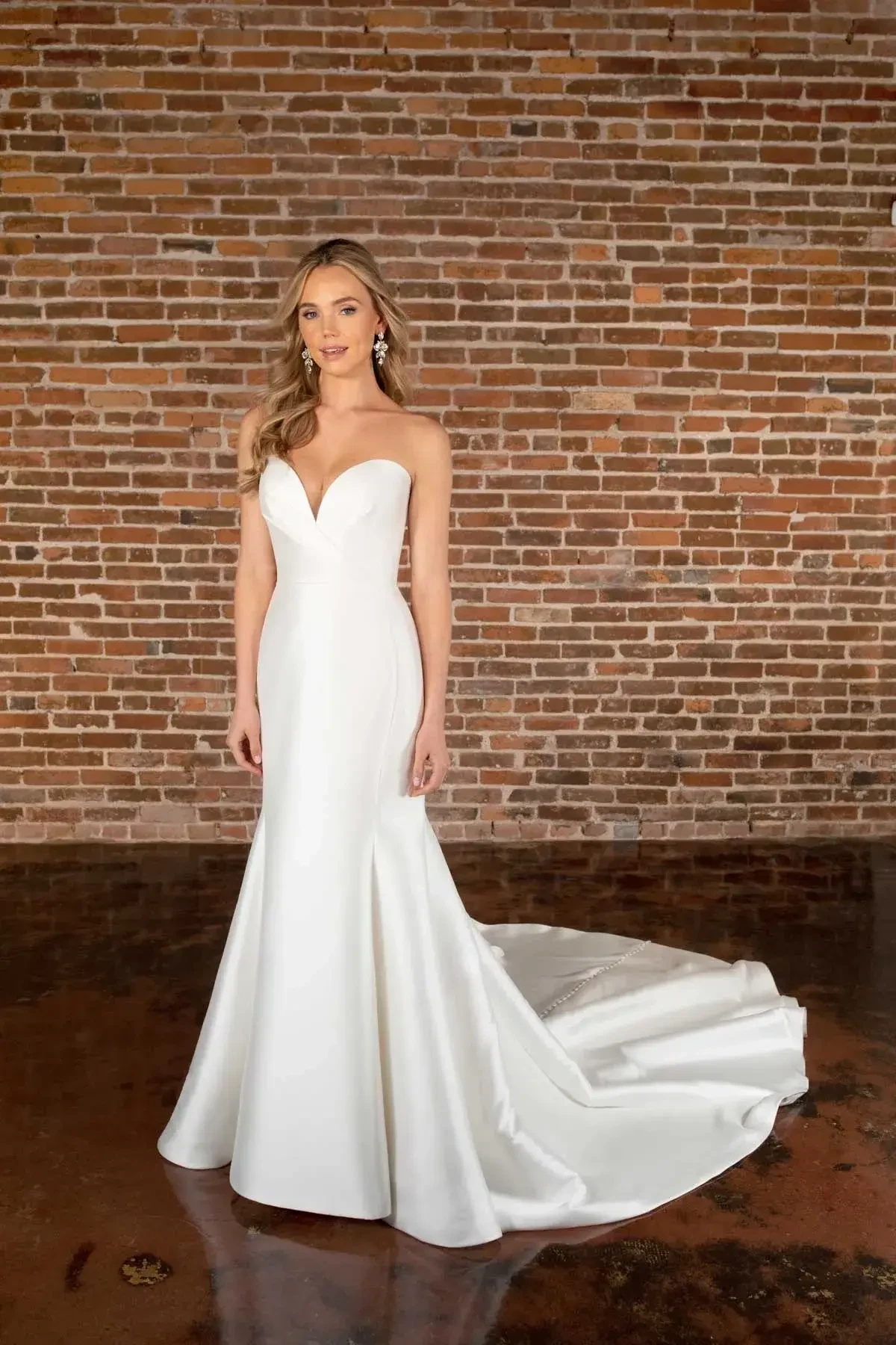 A woman in a strapless white wedding gown stands on a polished floor against a brick wall. Her expression is serene, conveying elegance and poise.