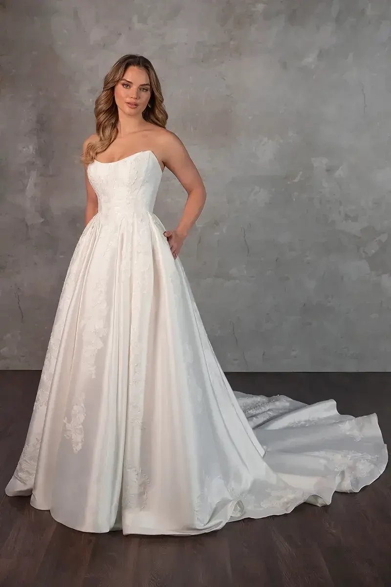 Model is wearing Style D4265 by Essense of Australia, a strapless Mikado ball gown with a scooped cat-eye neckline, basque waist, pleated skirt, dramatic train, and fabric-covered buttons down the back.