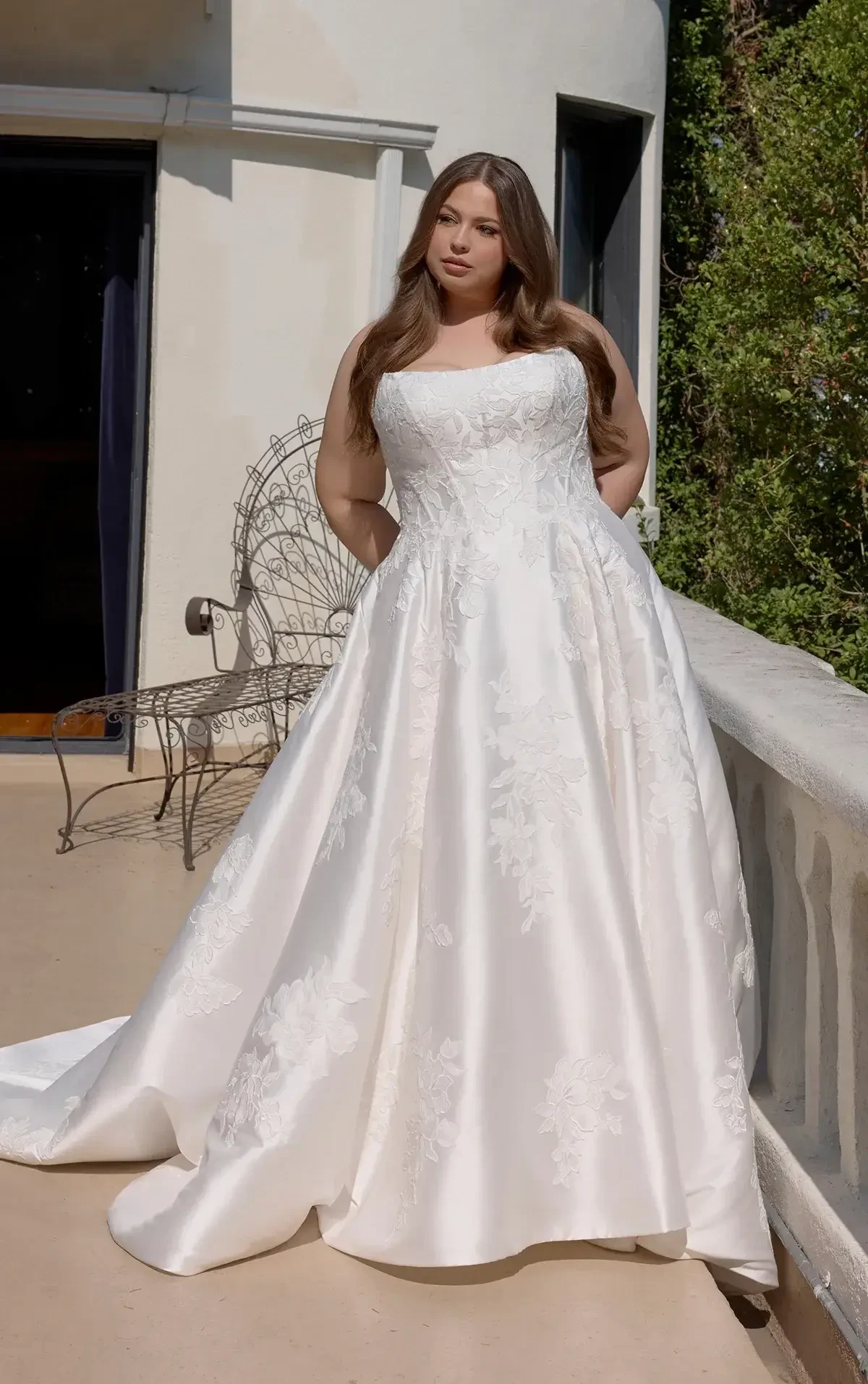 A woman in a white wedding dress stands on a balcony, looking confidently at the camera. The dress has a fitted bodice and a flowing skirt, with floral patterns visible on the fabric. Surrounding greenery can be seen in the background.