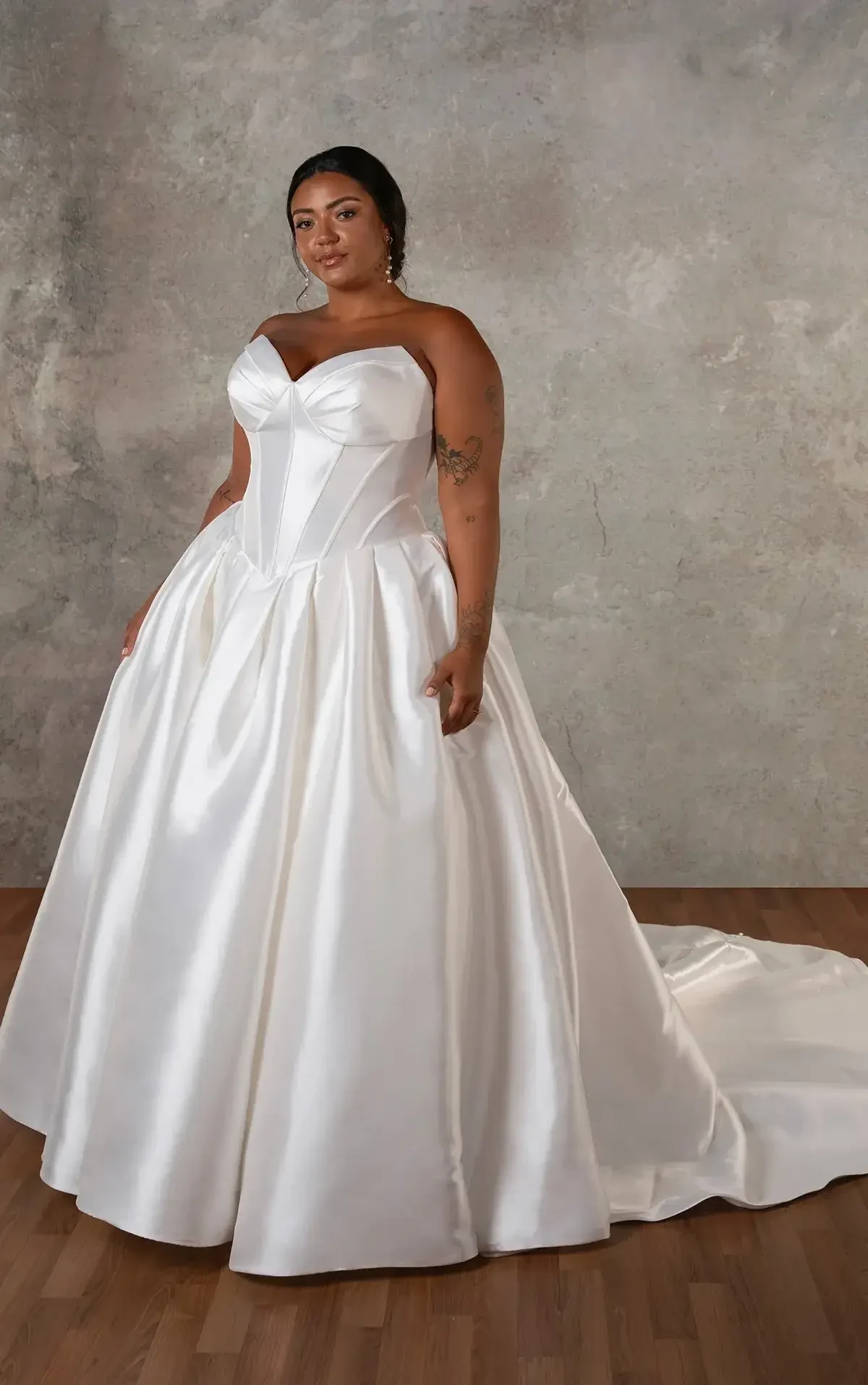A plus-size woman wearing a strapless, white satin wedding gown with a full skirt, standing against a textured gray background.
