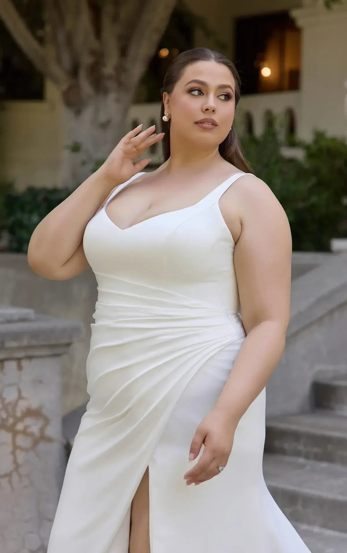 A woman in a white dress poses outdoors, showing a confident expression. The setting features lush greenery and stairs in the background.