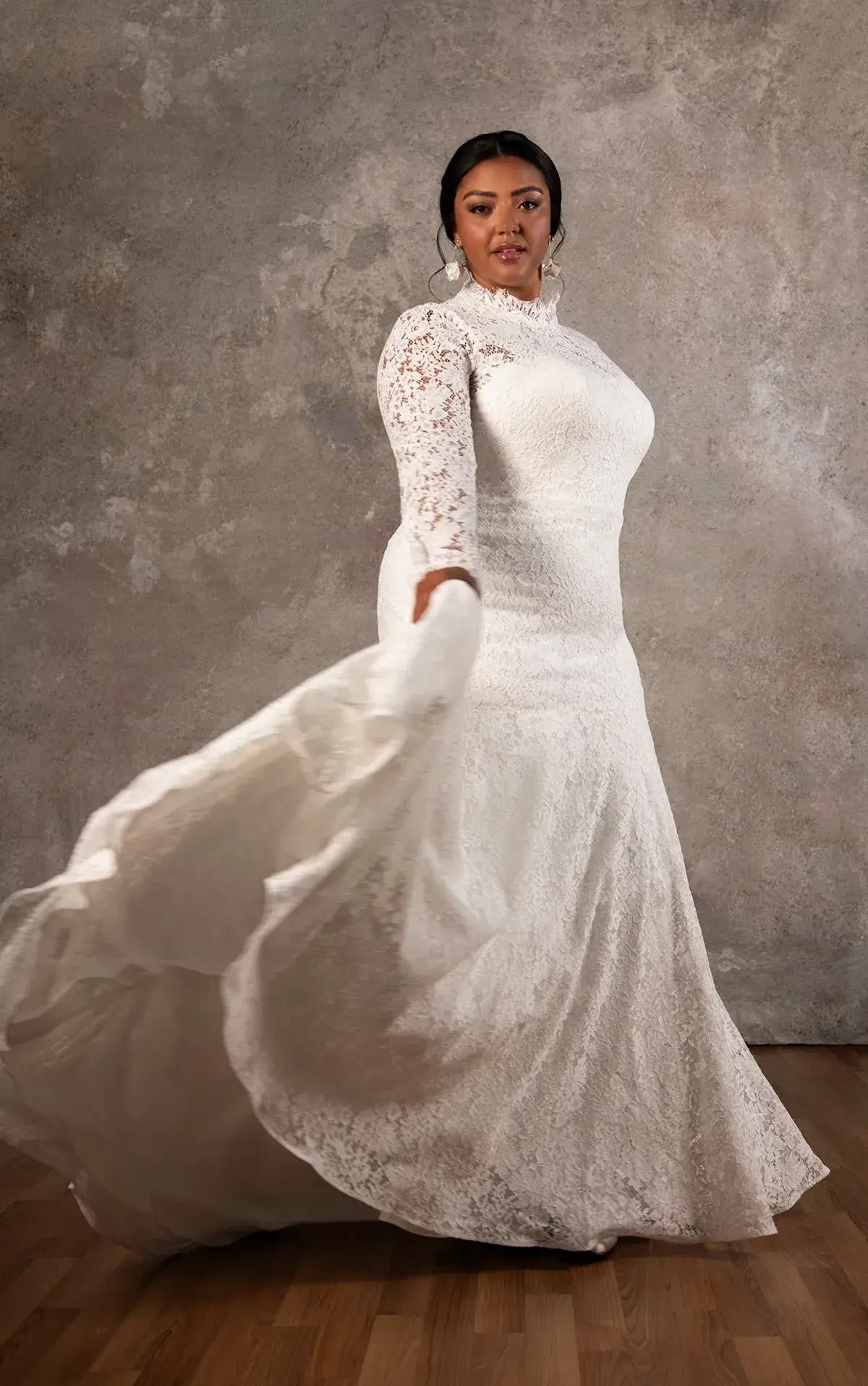 A woman in a white lace wedding dress poses gracefully, holding the flowing fabric of her gown. The background is a textured gray, and the lighting highlights her features and the intricate lace design of the dress.
