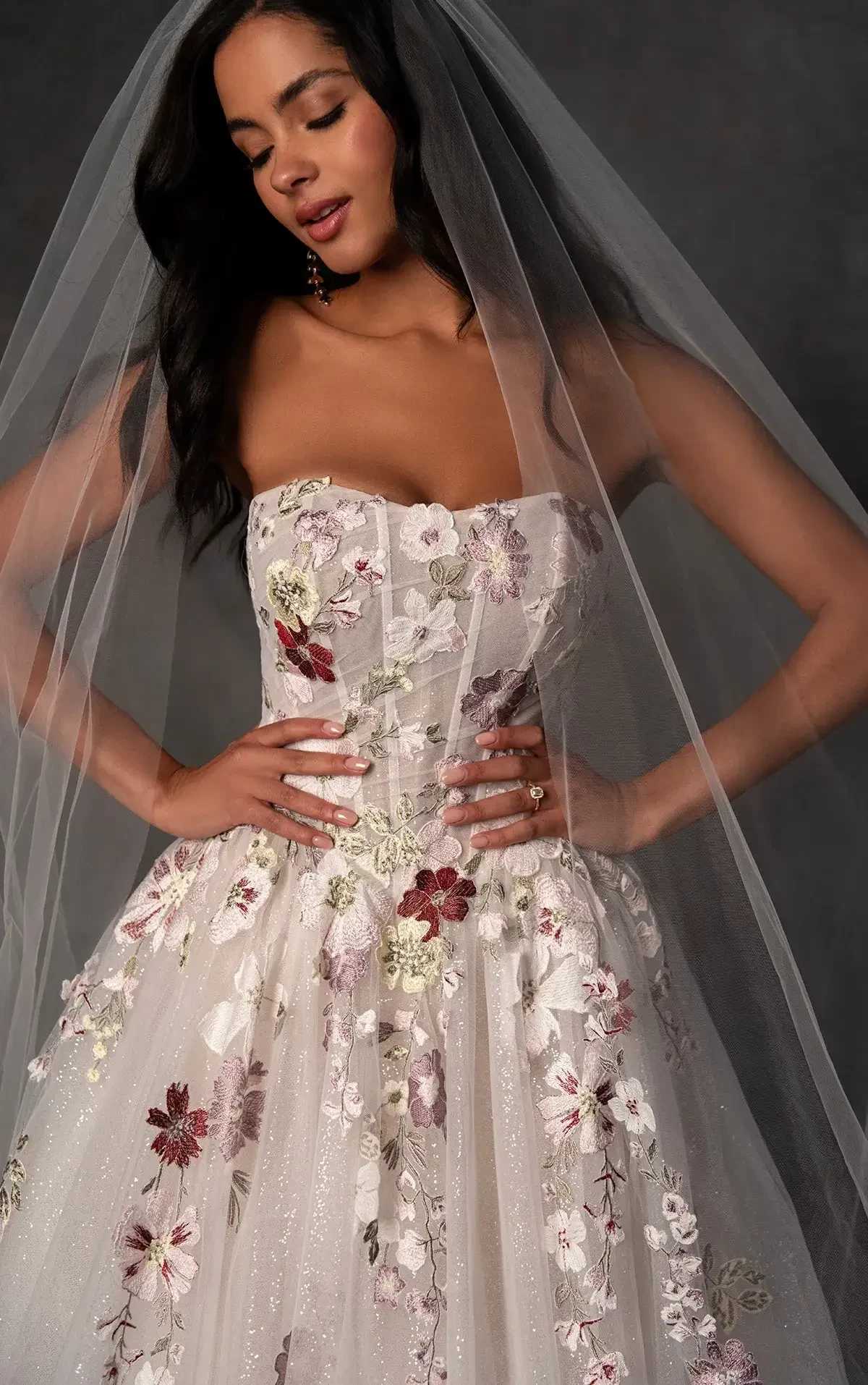 A bride in a strapless floral gown with intricate embroidery and a sheer veil stands confidently, smiling gently, evoking joy and elegance.