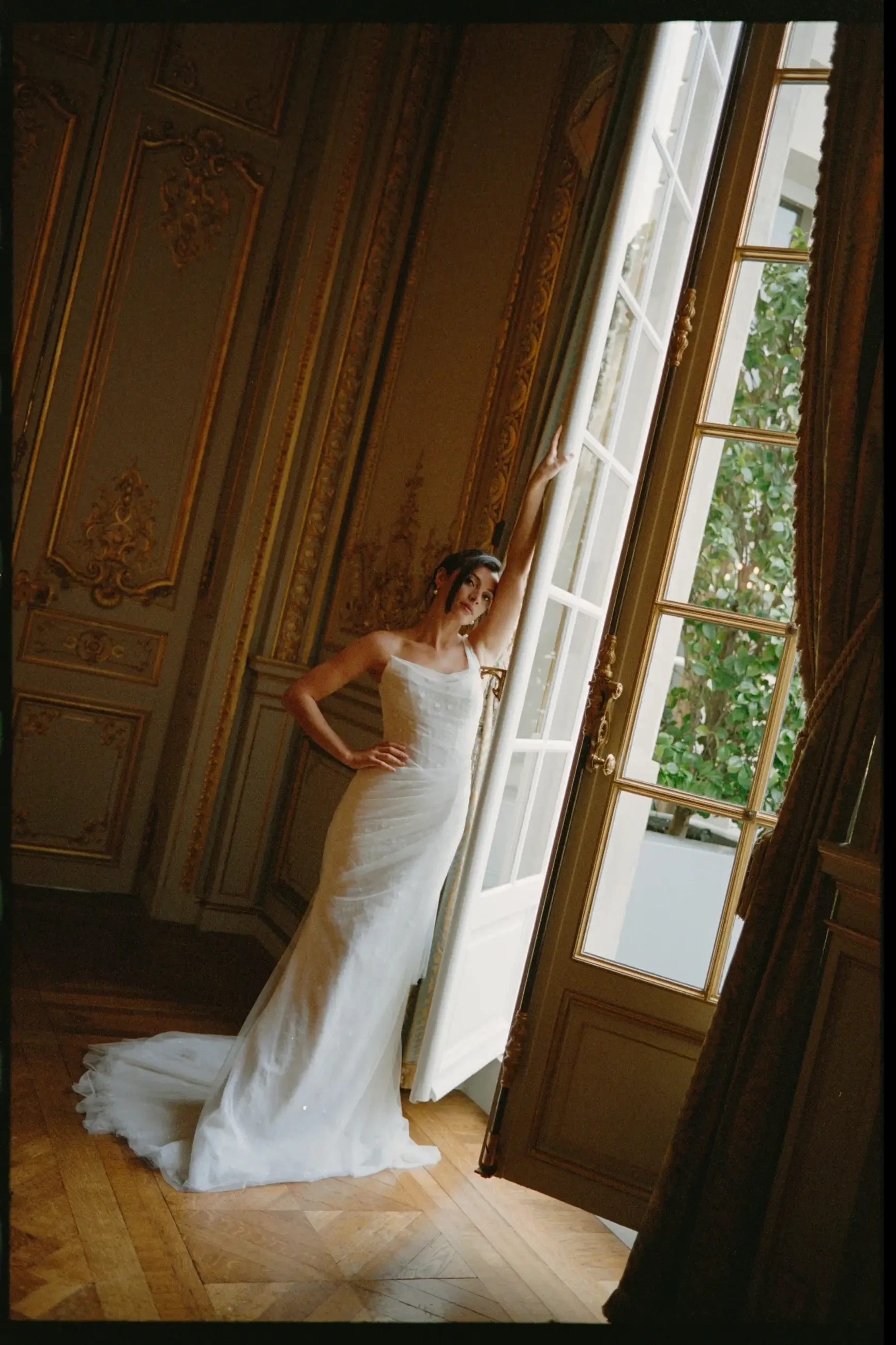 A bride in a white wedding dress poses by an open door, showcasing elegant interior decor with golden accents.