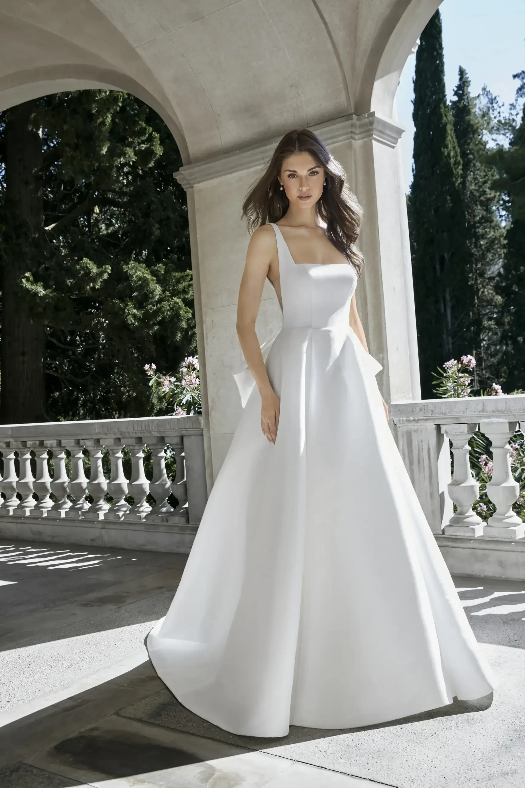 Model is wearing the Gigi gown by Kelly Faetanini, crafted in stretch Mikado with a square neckline, inverse box pleat skirt, and a detachable oversized bow with optional streamers.