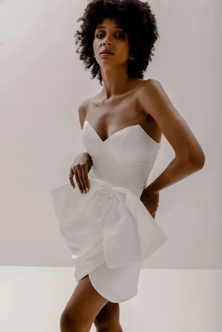 A model wearing a strapless white dress with a large bow detail, posing against a plain background. The model has curly hair and is looking confidently at the camera.