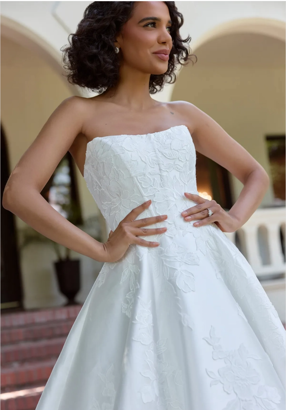 A woman with curly hair wearing a strapless white wedding dress, standing confidently with her hands on her hips.