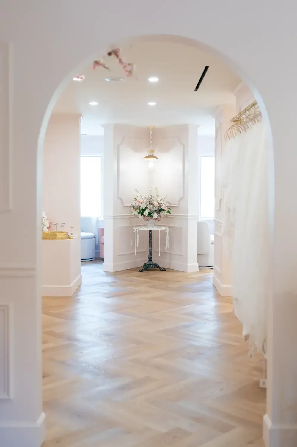 A well-lit hallway with a rounded arch, featuring light hardwood flooring and white walls. There is a small table with flowers at the end of the hall, and soft furnishings visible in the background.