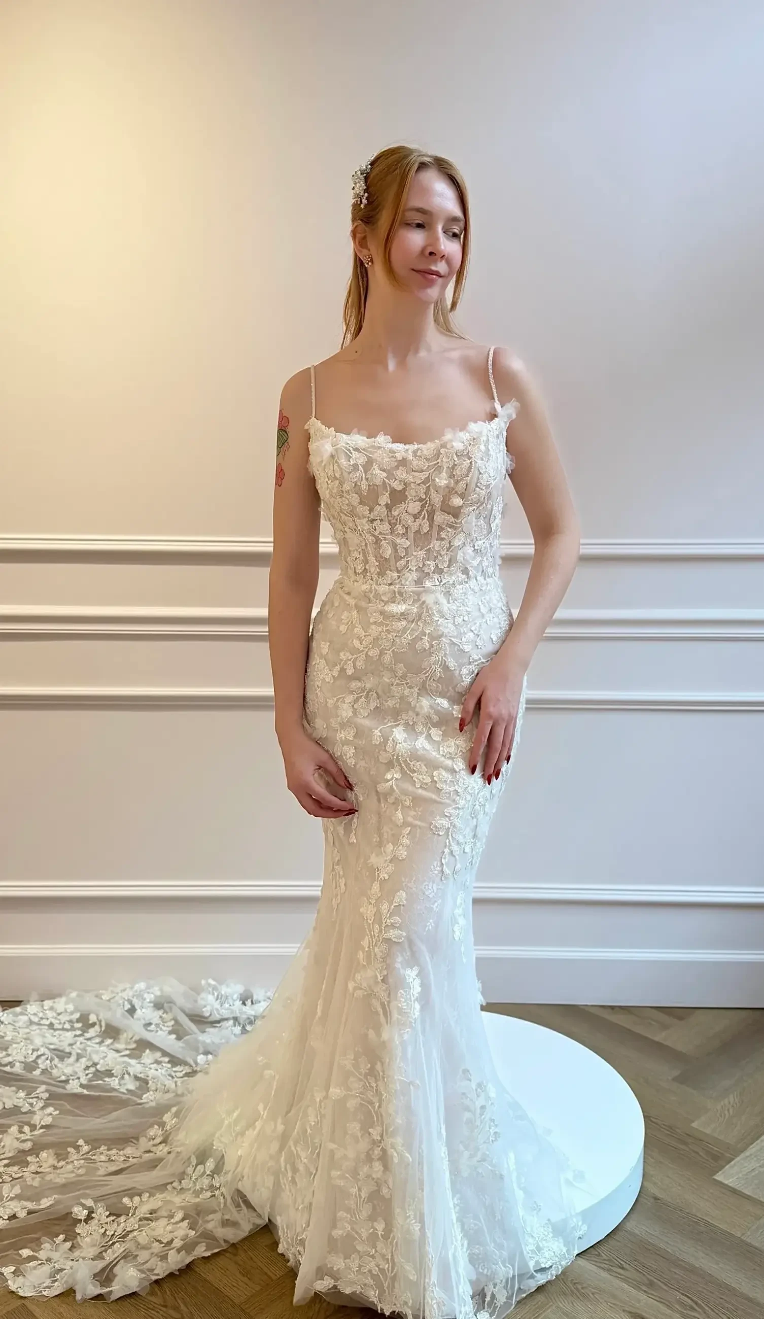 A woman wearing an intricately designed white bridal gown with floral details, posing in a bright, elegantly decorated room.