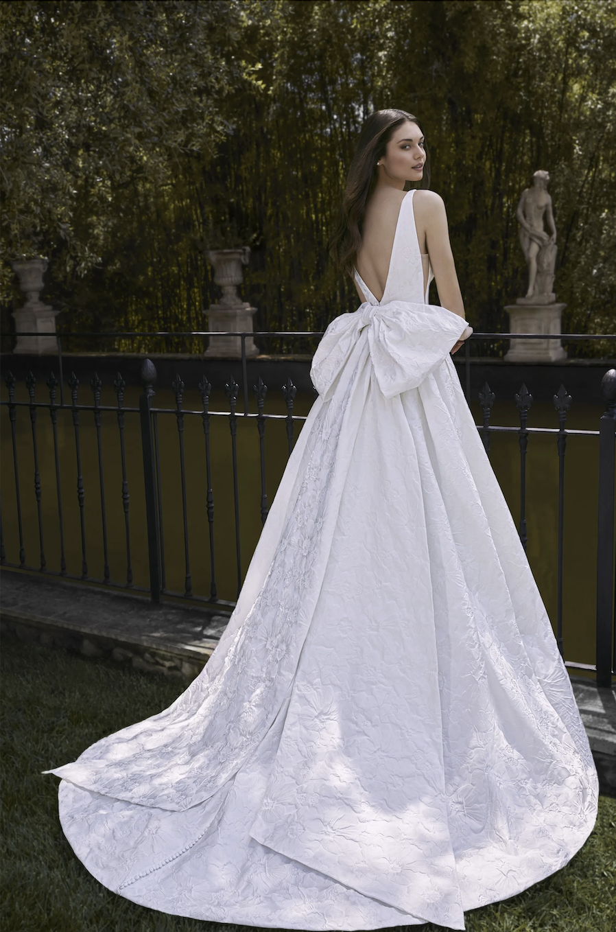 Model is wearing the Gigi gown by Kelly Faetanini, crafted in stretch Mikado with a square neckline, inverse box pleat skirt, and a detachable oversized bow with optional streamers.