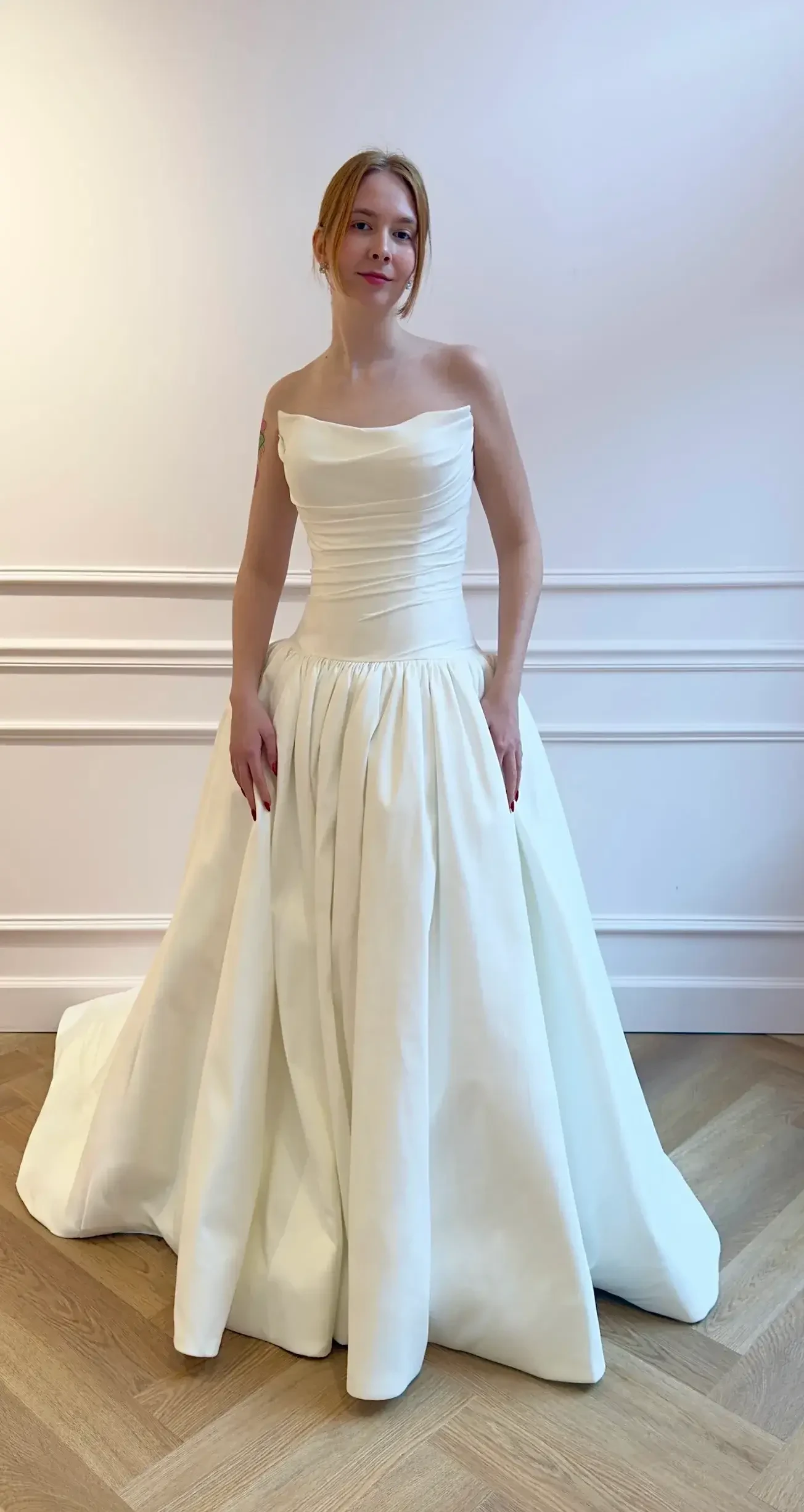 A woman in a strapless white gown stands in an elegantly decorated room with white walls and wooden flooring.