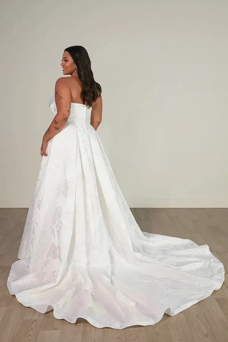Model is wearing SY8084 by Stella York, a strapless ball gown with a ruched neckline, basque waist, pleated grand satin skirt, and a gown-length row of fabric-covered buttons.