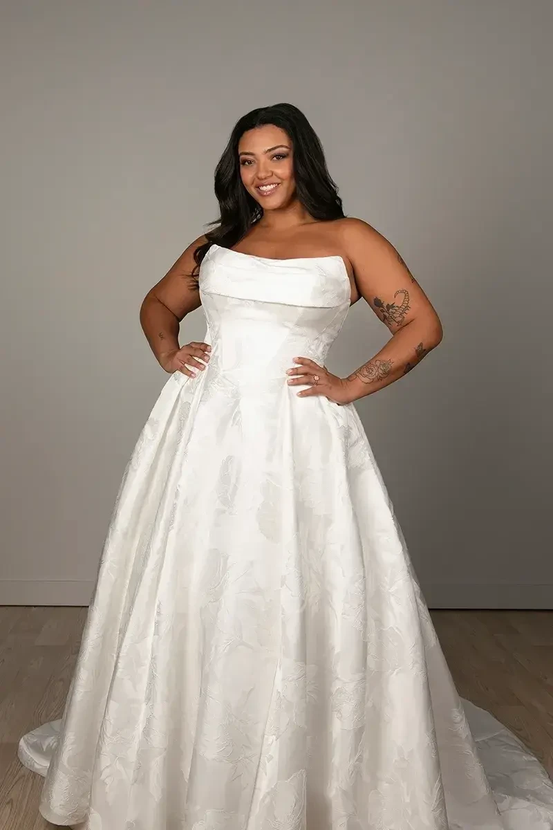 Model is wearing SY8084 by Stella York, a strapless ball gown with a ruched neckline, basque waist, pleated grand satin skirt, and a gown-length row of fabric-covered buttons.