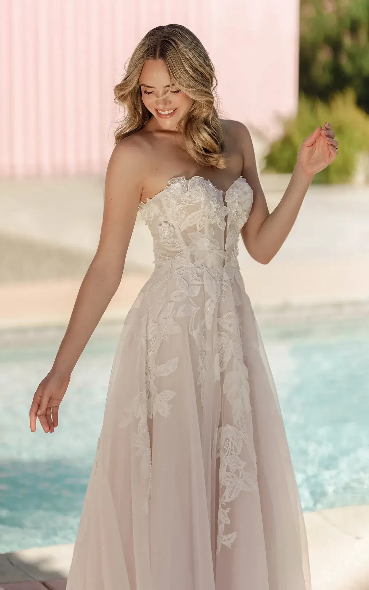 A smiling woman in a strapless, floral lace wedding dress stands by a pool. Her blonde hair cascades in waves, conveying a joyful, elegant vibe.