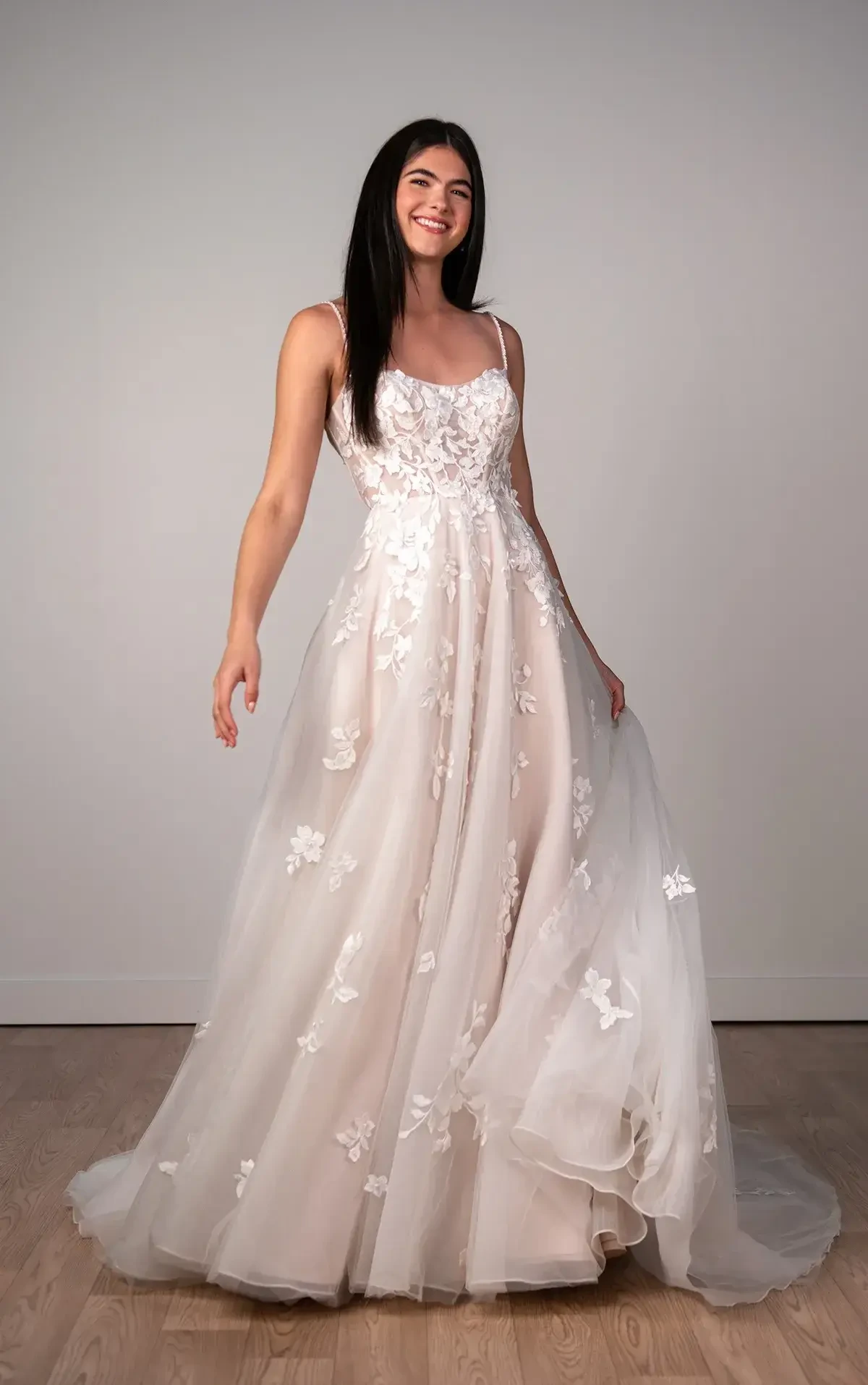 Smiling woman in an elegant white wedding gown with floral lace details and a flowing skirt, stands on wooden floor against a gray background.