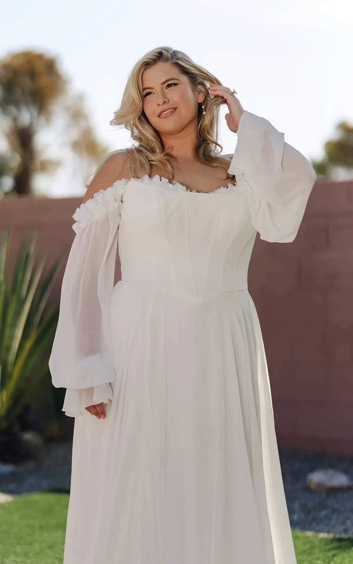 A woman in a flowing white off-shoulder gown stands outside, smiling thoughtfully. Greenery and a pink wall backdrop add a serene tone to the setting.